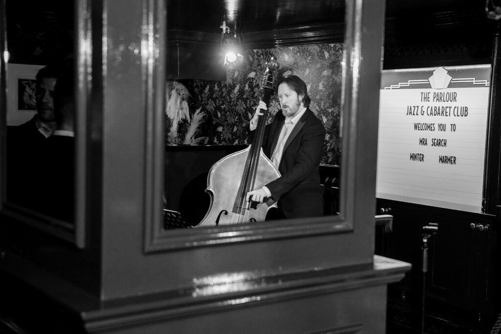 A photograph showing a reflection of a man playing the double bass, with a sign in the background.